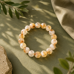 Unheated Citrine 10mm bracelet made of round translucent white and pale yellow gemstone beads with orange-brown inclusions, arranged in a circle on an olive green fabric surface with leaf and stone props. The brand name The Zen Crystals appears near the top.