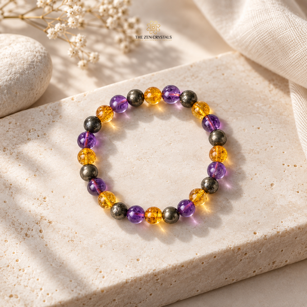 A beaded stretch bracelet by The ZenCrystals, featuring translucent purple amethyst and golden-yellow citrine beads alongside matte dark gray/black metal beads, displayed on a textured beige stone slab.