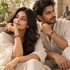 A man and a woman in cream-colored traditional clothing sit close together outdoors, each wearing a beaded bracelet with purple, yellow, and blue beads. The woman wears a gold ring on her finger.
