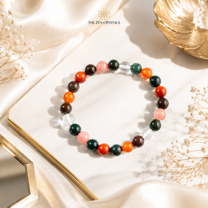 Aries zodiac bracelet arranged in a circle on a white marble slab with gold trim, surrounded by draped satin fabric, dried flowers, and a gold decorative object. The bracelet features round polished beads in orange, dark green, pink, brown, and clear.