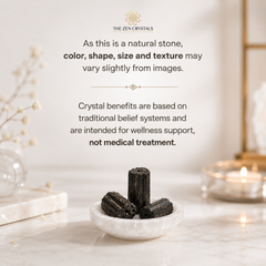 Three dark natural stone cylindrical pieces displayed in a small white bowl on a marble surface, with soft-focus decor and a candle in the background. Text at the top reads 'THE ZEN CRYSTALS' and includes a disclaimer about color, shape, size, and texture varying slightly from images.