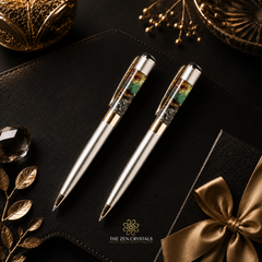 Two money magnet ballpoint pens with clear inlay sections showing a landscape design, presented on a black leather surface with decorative gold-toned props and a gift box with a satin bow. The Zen Crystals logo is visible at the bottom.