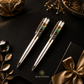 Two money magnet ballpoint pens with clear inlay sections showing a landscape design, presented on a black leather surface with decorative gold-toned props and a gift box with a satin bow. The Zen Crystals logo is visible at the bottom.