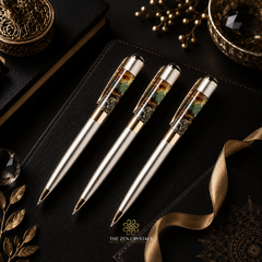 Three luxury money magnet ballpoint pens with metallic gold bodies and green-and-brown patterned resin accents in the clip areas, arranged on a black textured notebook. Gold decorative props, a ribbon, and a crystal are visible around them.