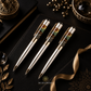 Three luxury money magnet ballpoint pens with metallic gold bodies and green-and-brown patterned resin accents in the clip areas, arranged on a black textured notebook. Gold decorative props, a ribbon, and a crystal are visible around them.