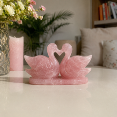 A rose quartz swan pair figurine on a matching base, placed on a glossy white table in a softly lit living room setting with a candle, flowers, and cushions in the background.