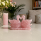 A rose quartz swan pair figurine on a matching base, placed on a glossy white table in a softly lit living room setting with a candle, flowers, and cushions in the background.