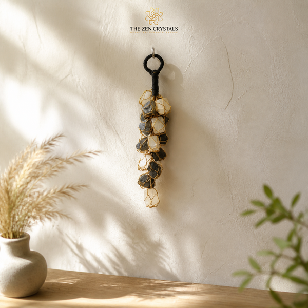 A decorative hanging wall ornament made of a black metal key-shaped top and a long strand of natural stones in beige, gray, and brown, suspended on a textured off-white wall above a wooden surface with dried grass and green leaves nearby.